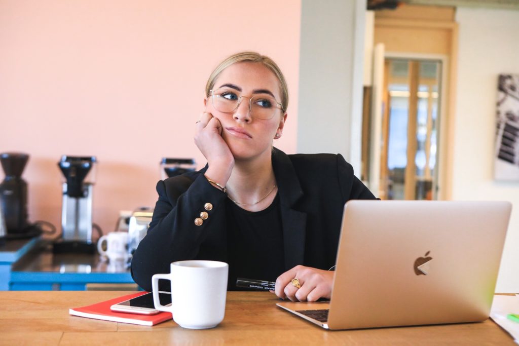 Eine Frau im Anzug sitzt an einem Tisch, vor Laptop und Kaffeetasse und denkt angestrengt-frustriert nach. Sie hat gerade erfahren, dass ihr ihr Nebenjob auf ALG 1 angerechnet wird. 