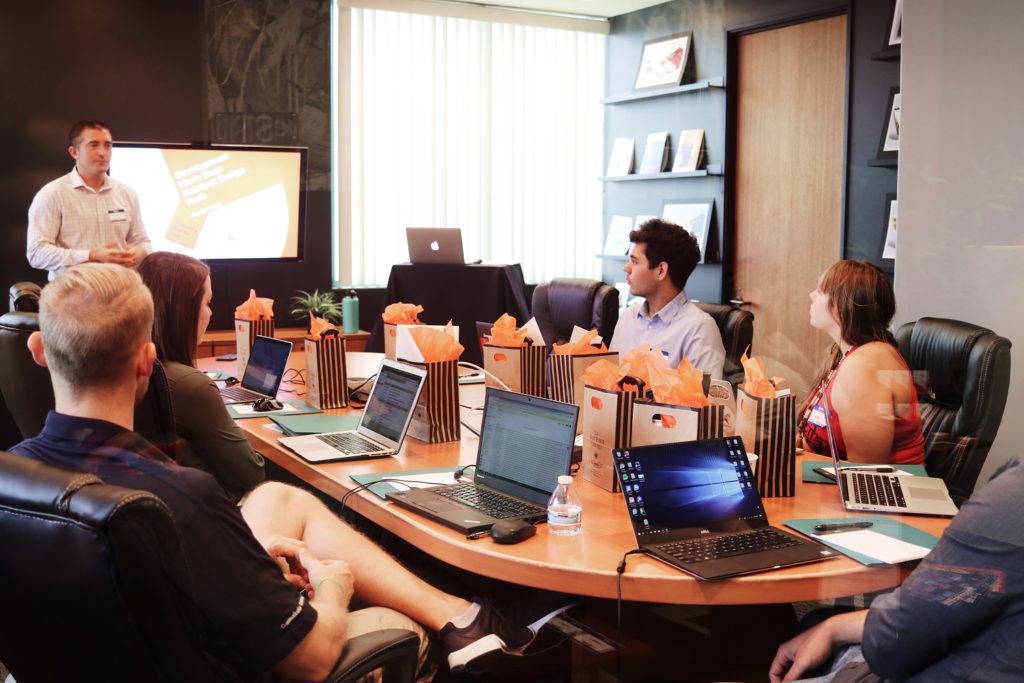 Ein Meeting mit mehreren Angestellten in einem Büro an einem länglichen Tisch. Ein Mitarbeiter steht vor dem Tisch und referiert.