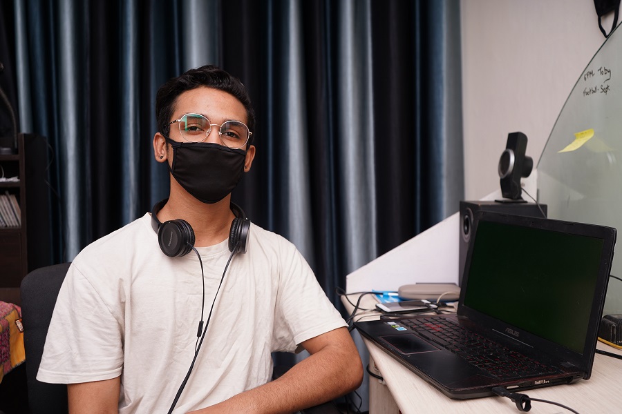 Ein junger Mann mit Kopfhörern um den Hals sitzt an einem Laptop in einem Büro. Er trägt eine Brille und einen Mund-Nase-Schutz und schaut in die Kamera. Er steht hier stellvertretend für Minijobber und Werkstudenten in der IT.