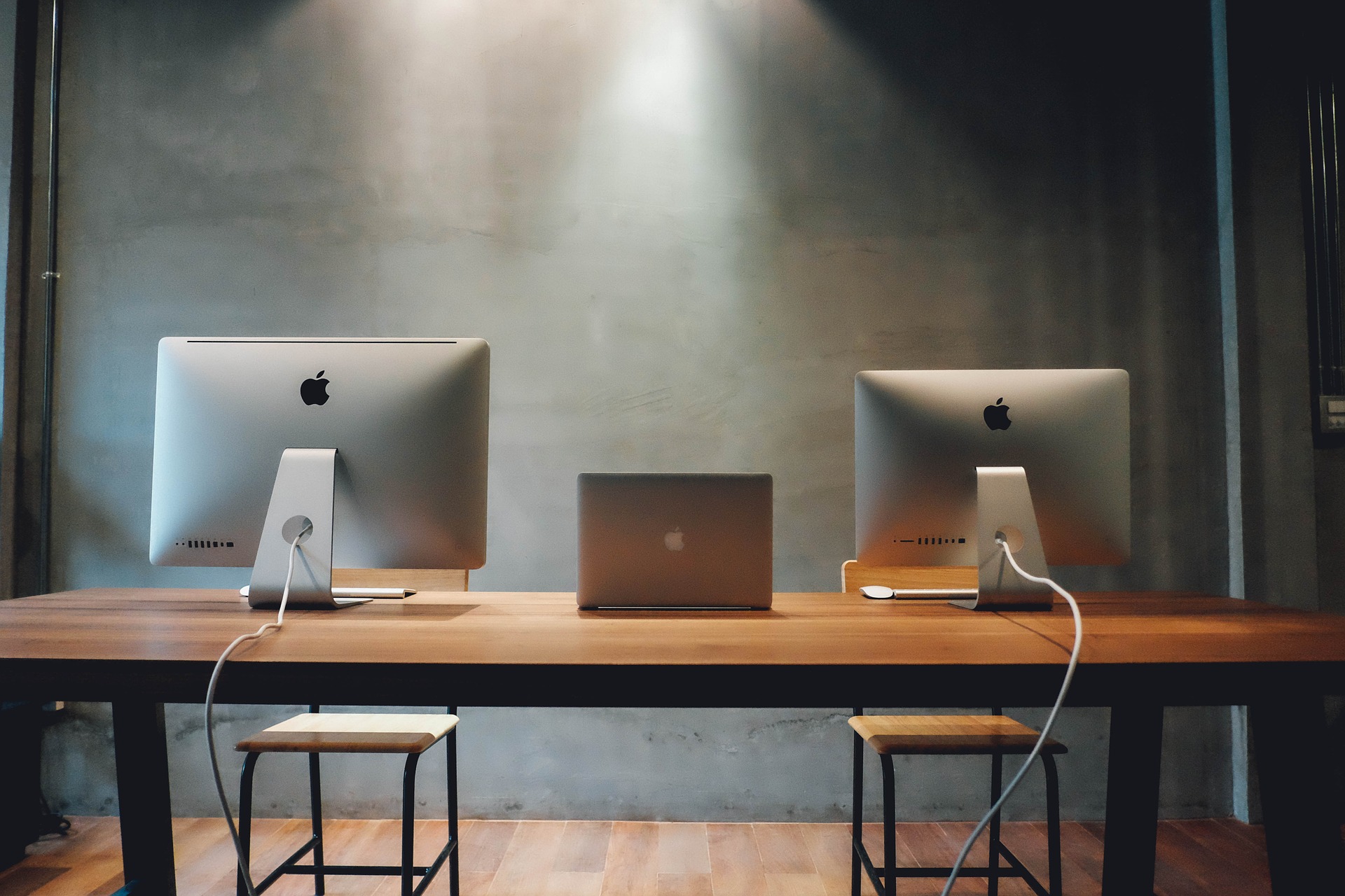 Zwei iMacs in verschiedenen Größen samt Maus und Tastatur sowie ein 15 Zoll MacBook Pro die beide auf einem Holztisch stehen. Die Holzstühle, die den Macs gegenüber platziert sind, stehen leer. Dies symbolisiert den Arbeitsplatz des Arbeitnehmers, der nach einer Kündigungsschutzklage nur selten in das Unternehmen zurückkehrt. 